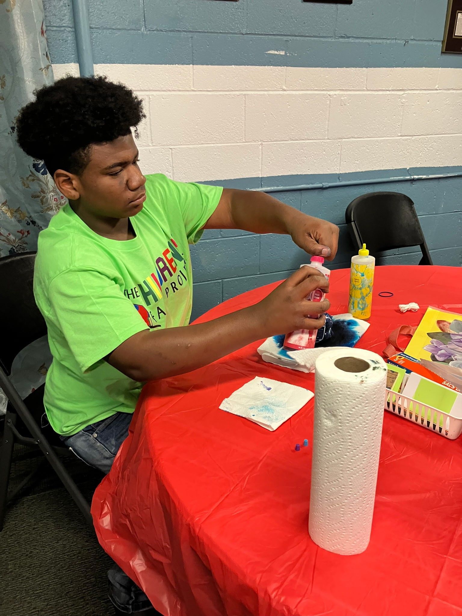 A person in a bright green shirt applying dye to fabric on a table covered by a red cloth.