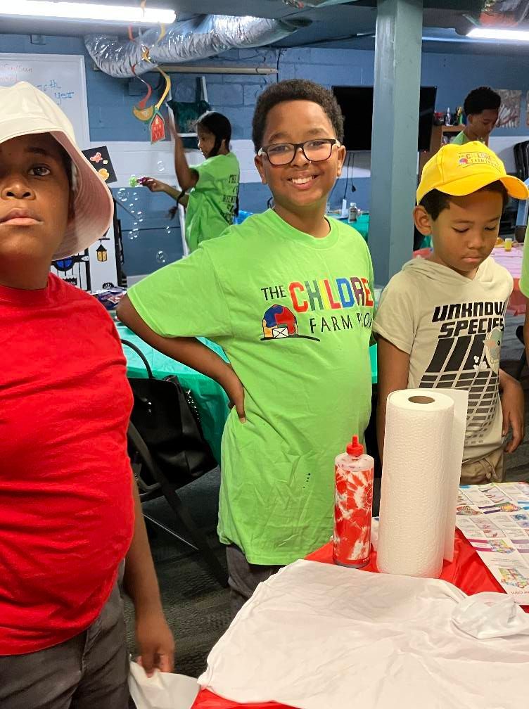 Three youth gathered around a craft table with supplies, one wearing a green t-shirt, in an indoor workspace.