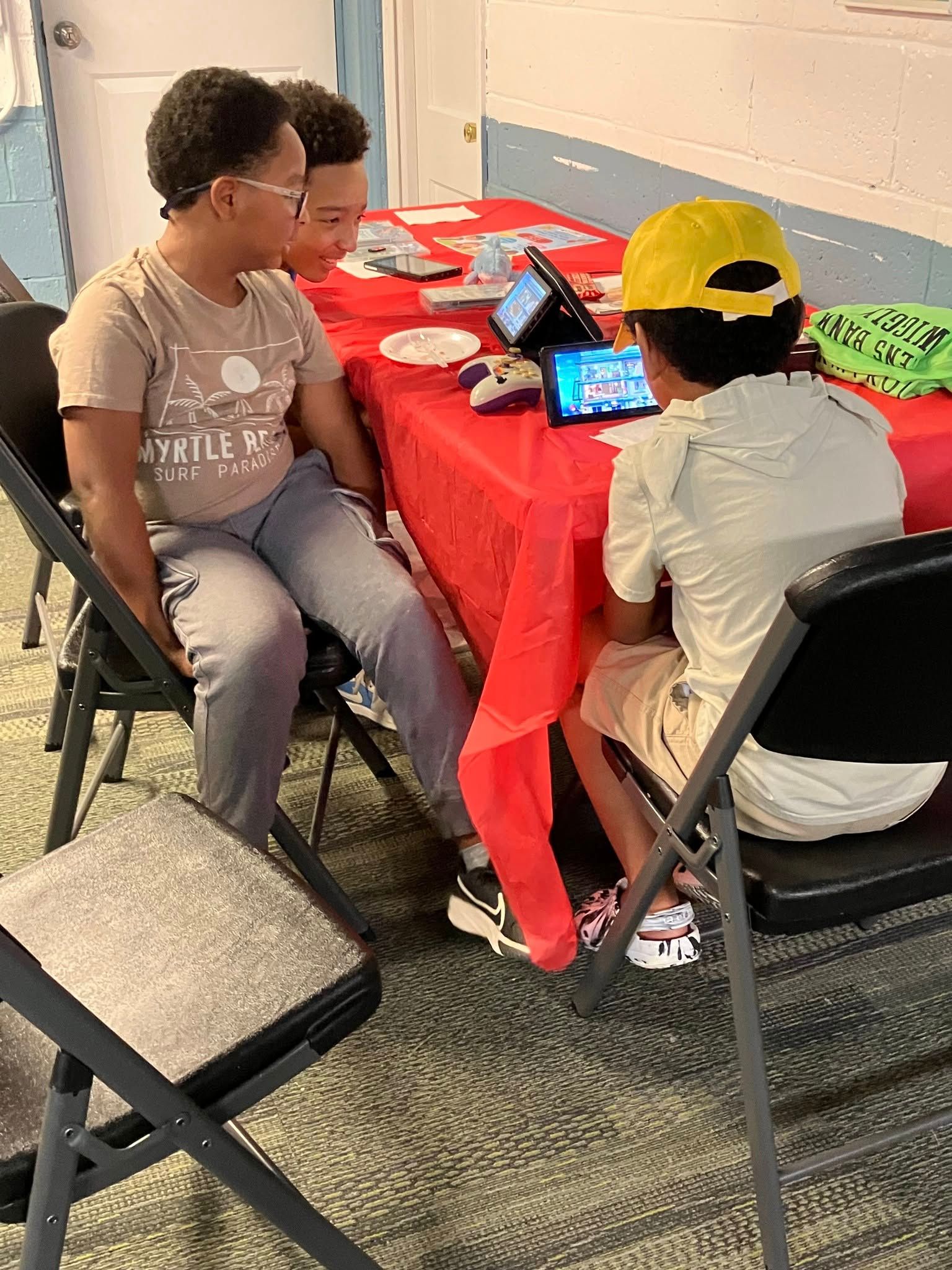Three children sit at a red-covered table playing video games on handheld devices inside a room with a block wall.