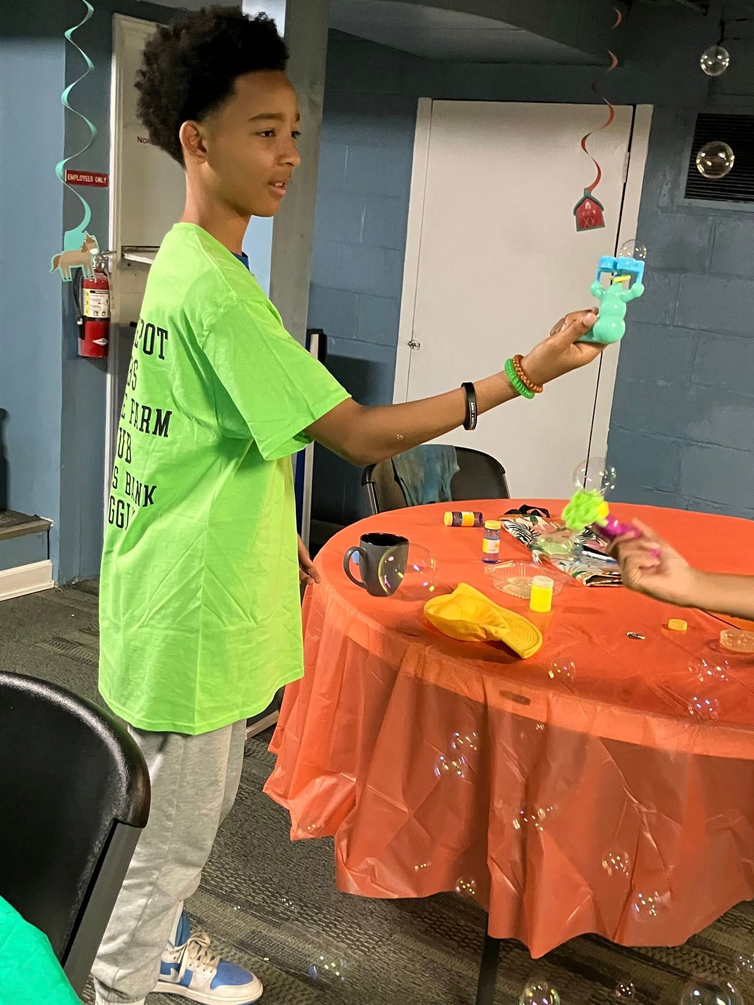 A person in a neon green shirt stands beside an orange-covered table, holding up a small teal toy.