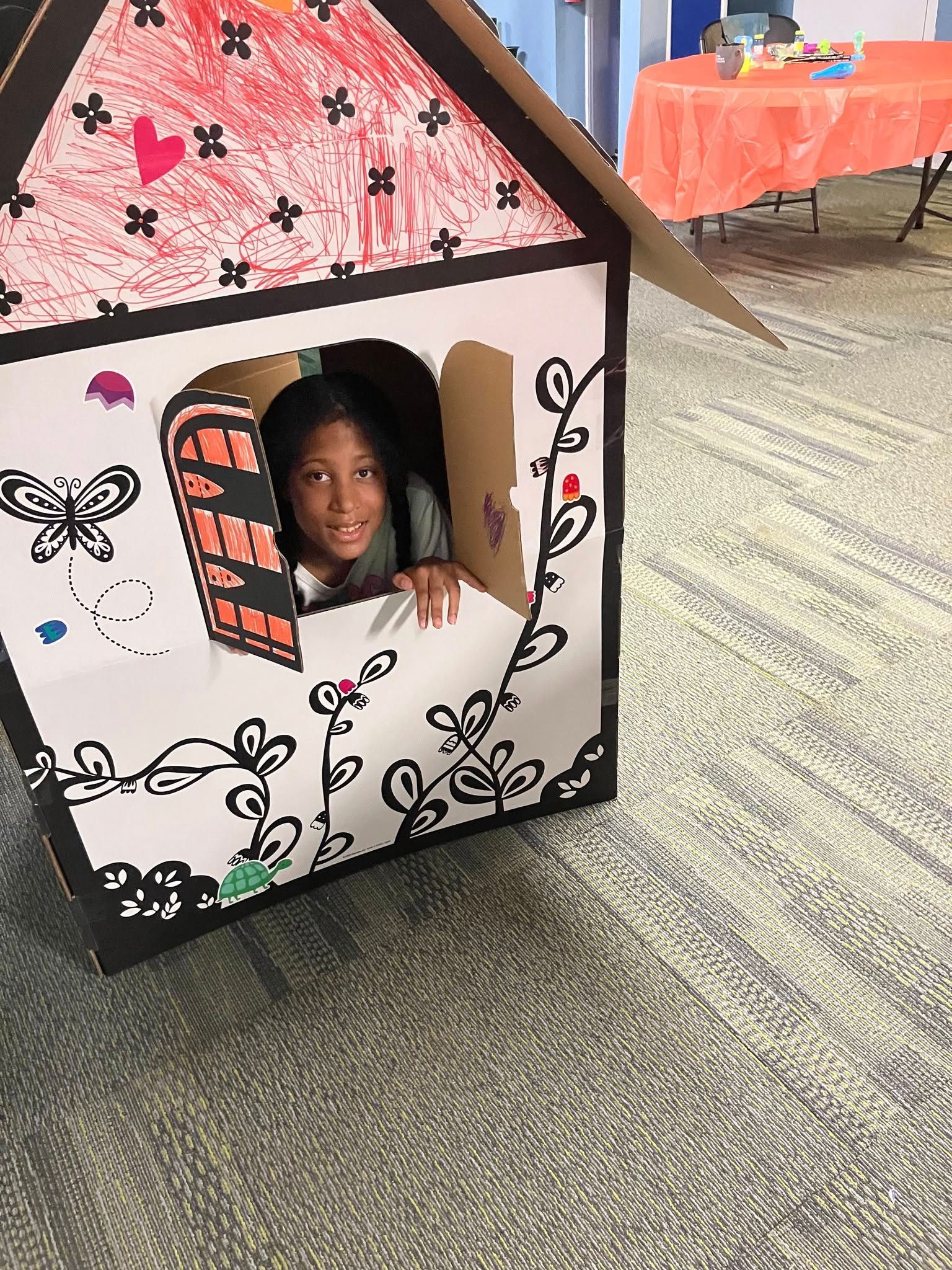 A person peers through a window of a decorated cardboard playhouse, situated on a carpeted floor near an orange table.