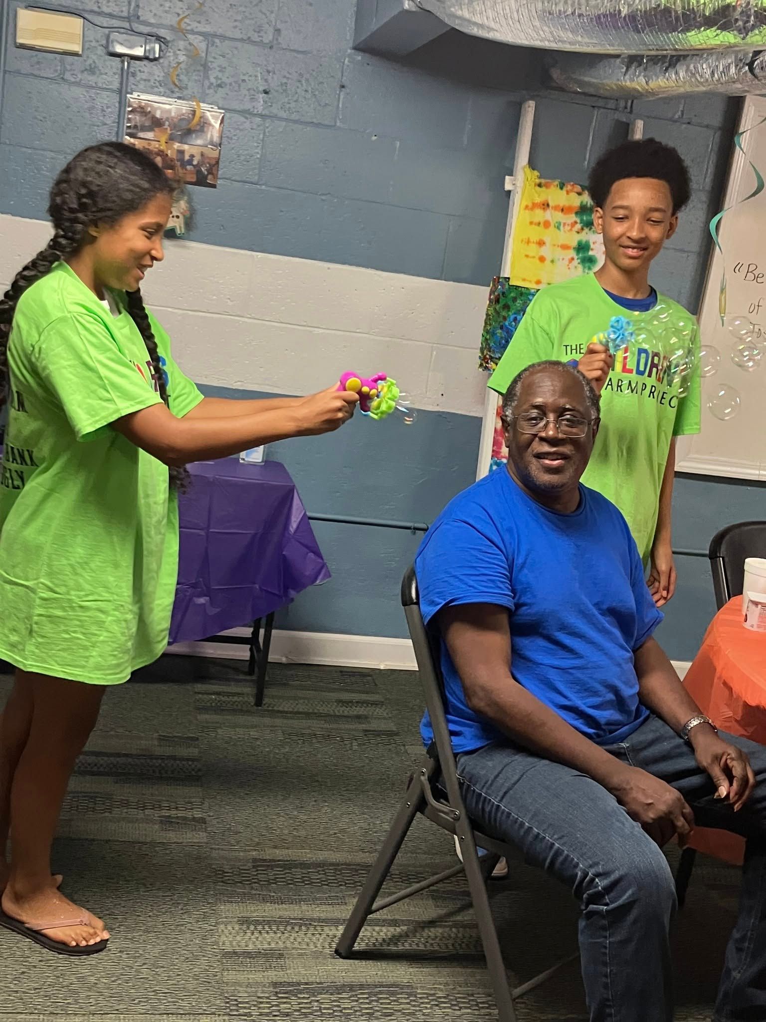 A young person in a green shirt holds a colorful toy toward a smiling adult in a blue shirt sitting on a chair.