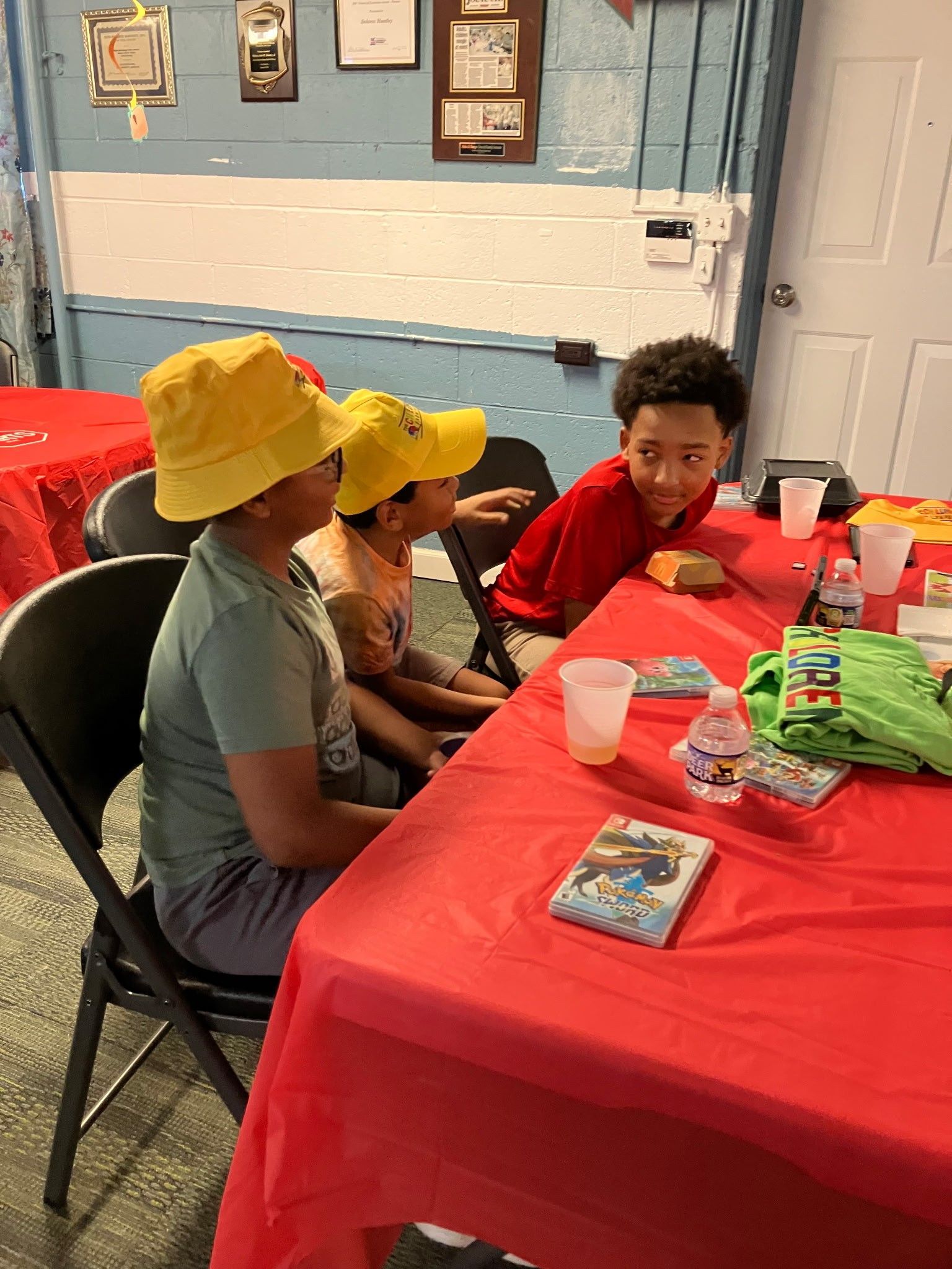 Three people seated at a table covered with a red tablecloth, two wearing yellow bucket hats, in a community room.