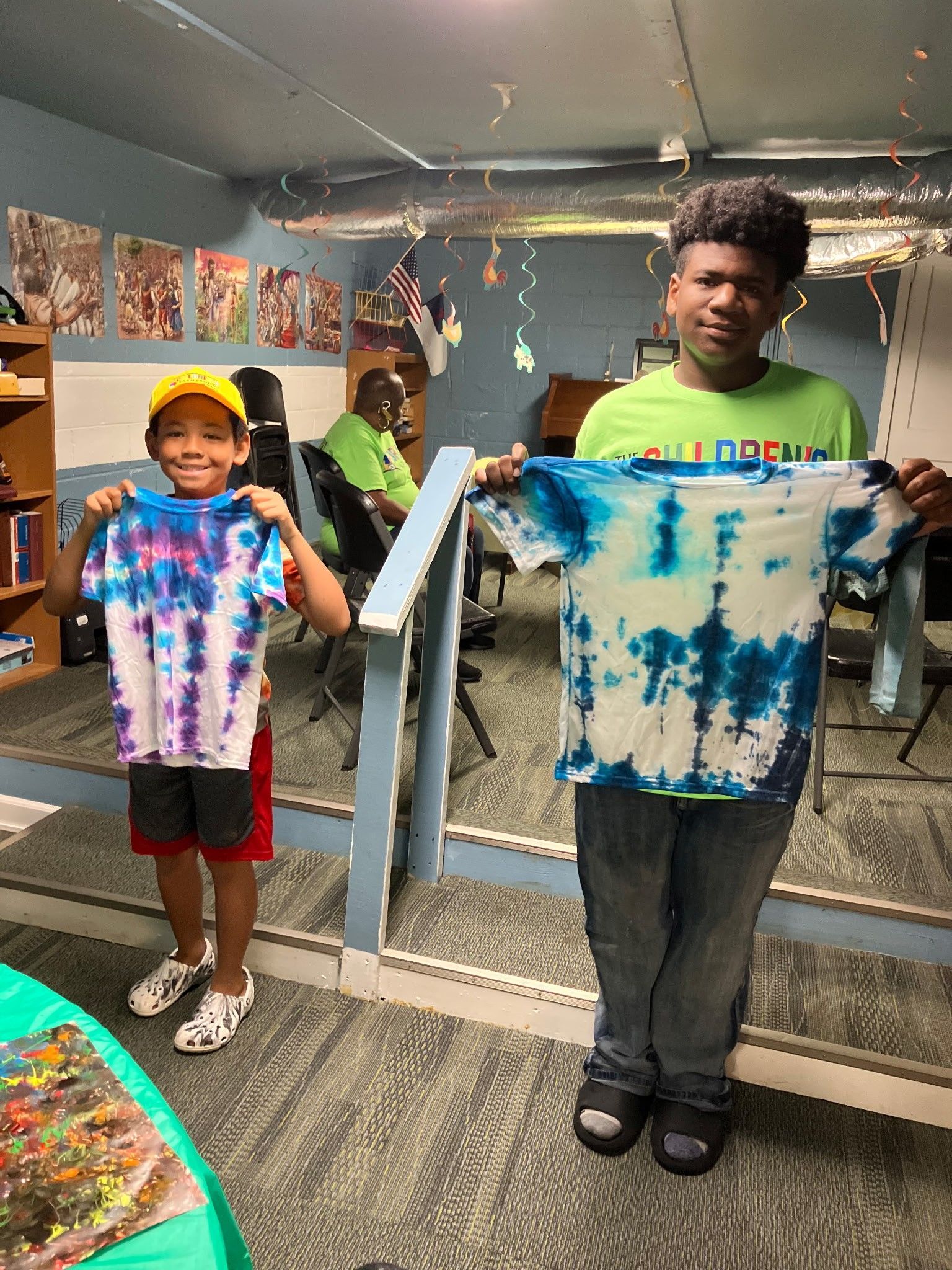 Two individuals hold up their handmade tie-dye shirts in a community room with patterned carpet and decorations.