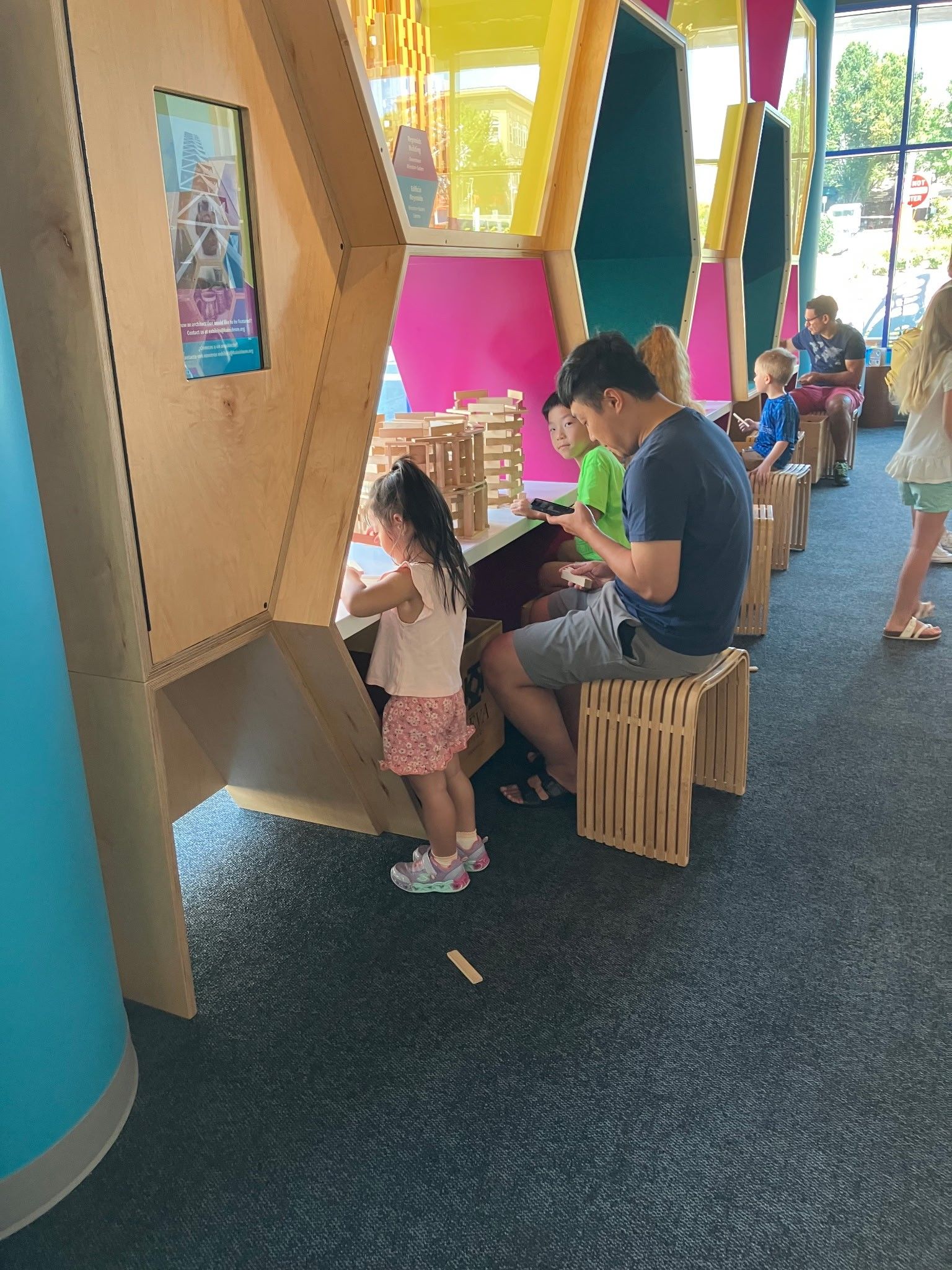 People sit at wooden honeycomb-shaped stations in a brightly colored indoor play area, focused on building with sticks.