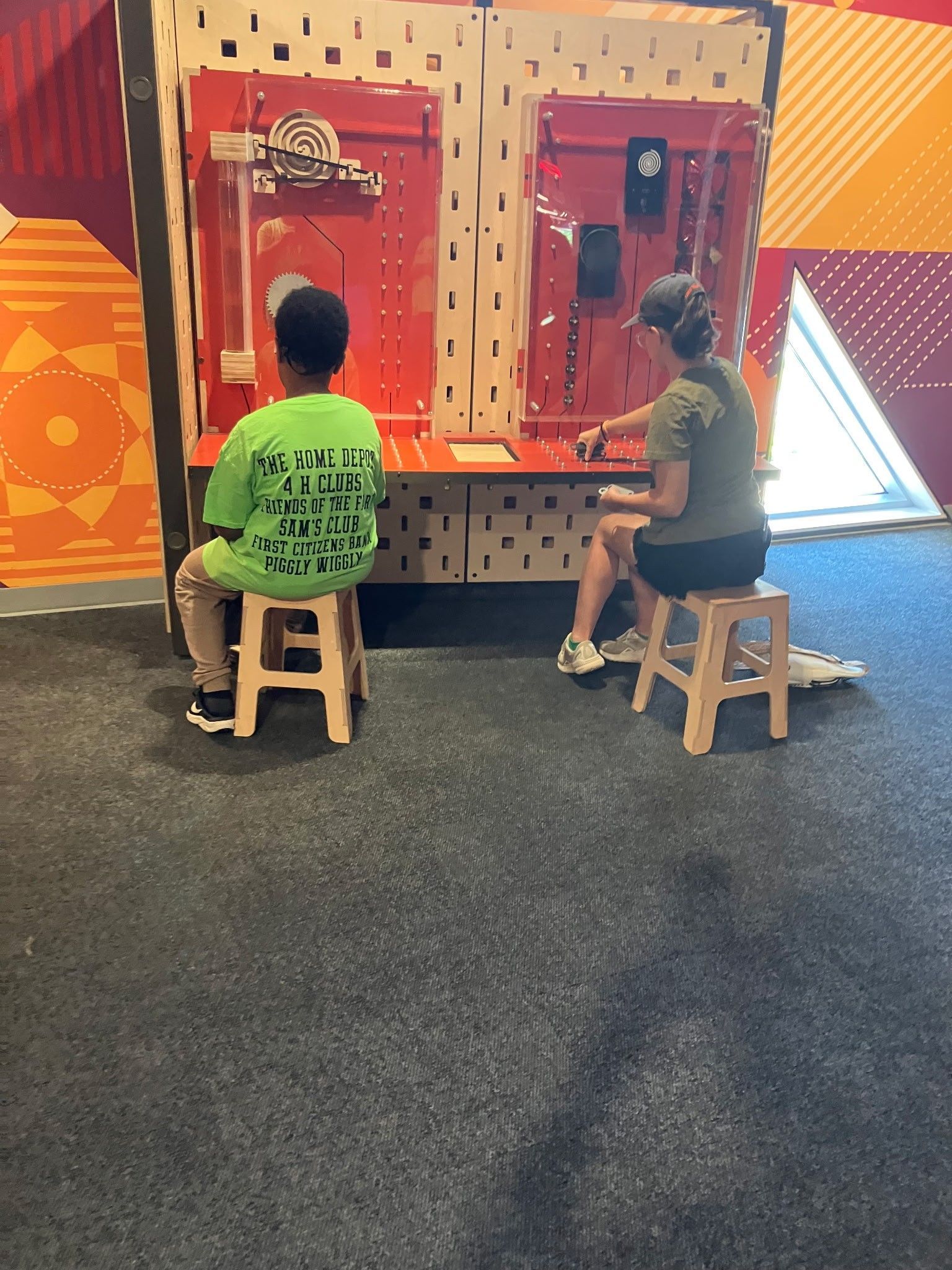 Two people sit on wooden stools at a wall-mounted activity board with red panels in a colorful room.