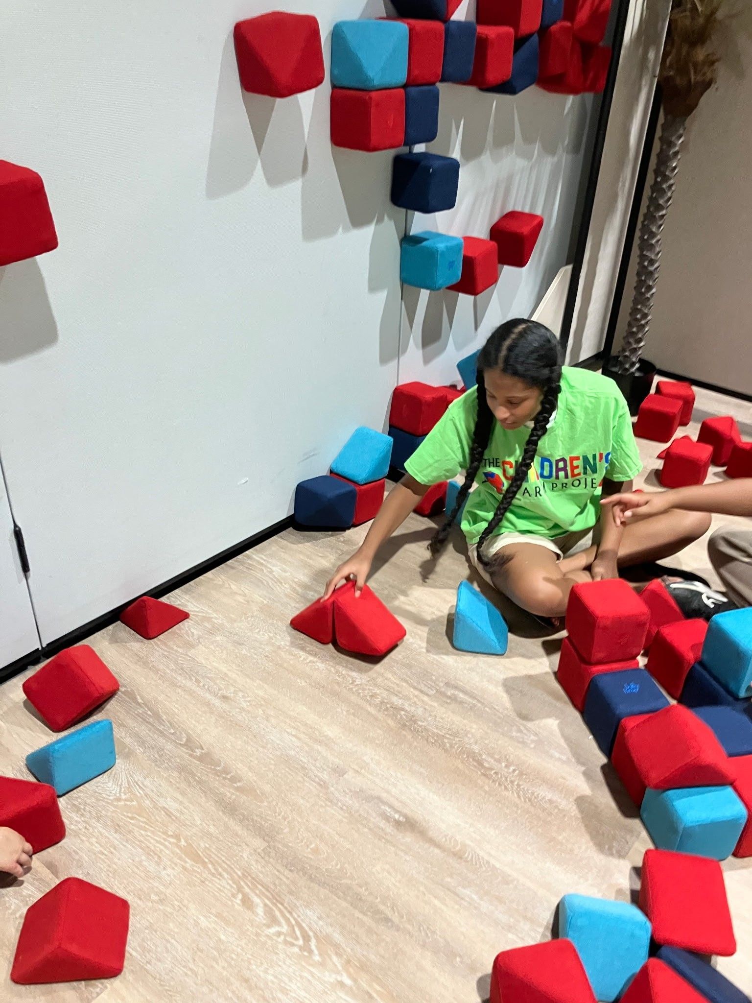 A person in a bright green shirt plays with red and blue foam building blocks on a light wood floor near a wall.