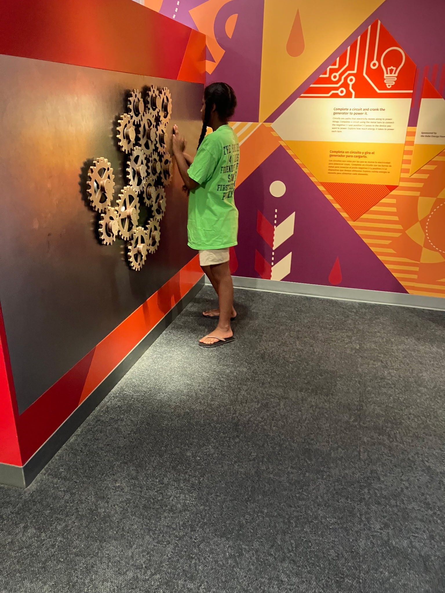 A person in a green shirt interacts with a wall-mounted gear puzzle in a brightly colored museum gallery.