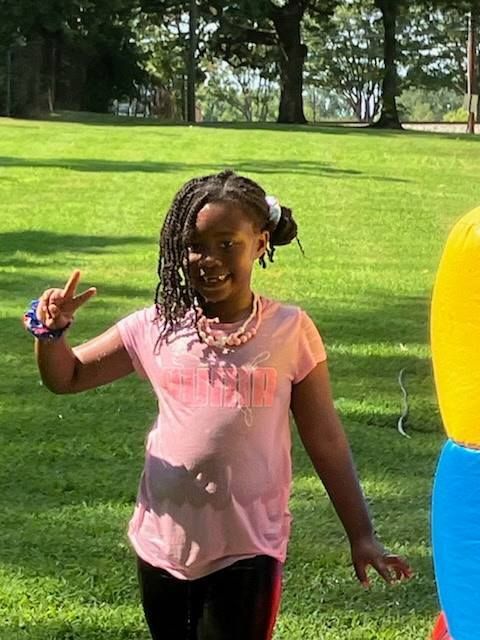 A person smiling and making a peace sign, wearing a light pink shirt and necklace, standing outside on a grassy lawn.