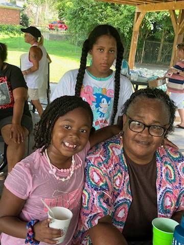 A smiling child and an older adult sit together at an outdoor gathering, with a youth standing behind them.