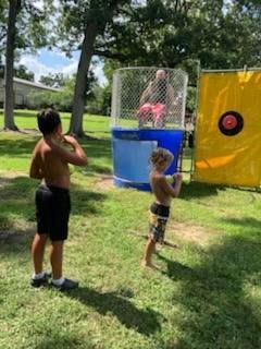 Two children prepare to throw a ball at a target to drop a person sitting in a blue water dunk tank outdoors.