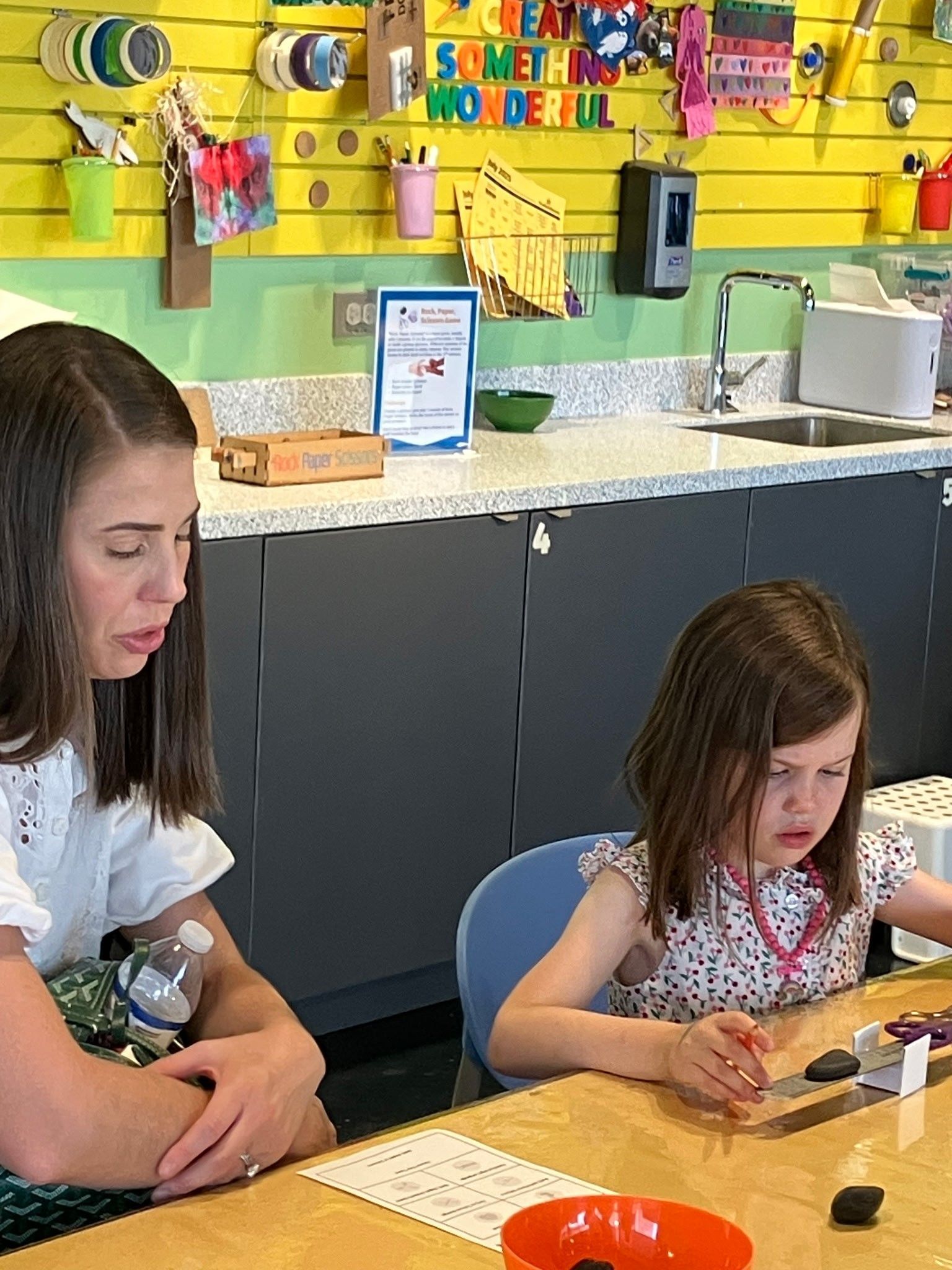A person looks on as a child sits at a table and plays with small objects in a colorful, well-lit craft room.