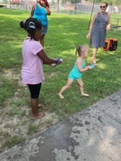 Two children play with water toys on a grassy lawn while two adults stand in the background on a sunny day.