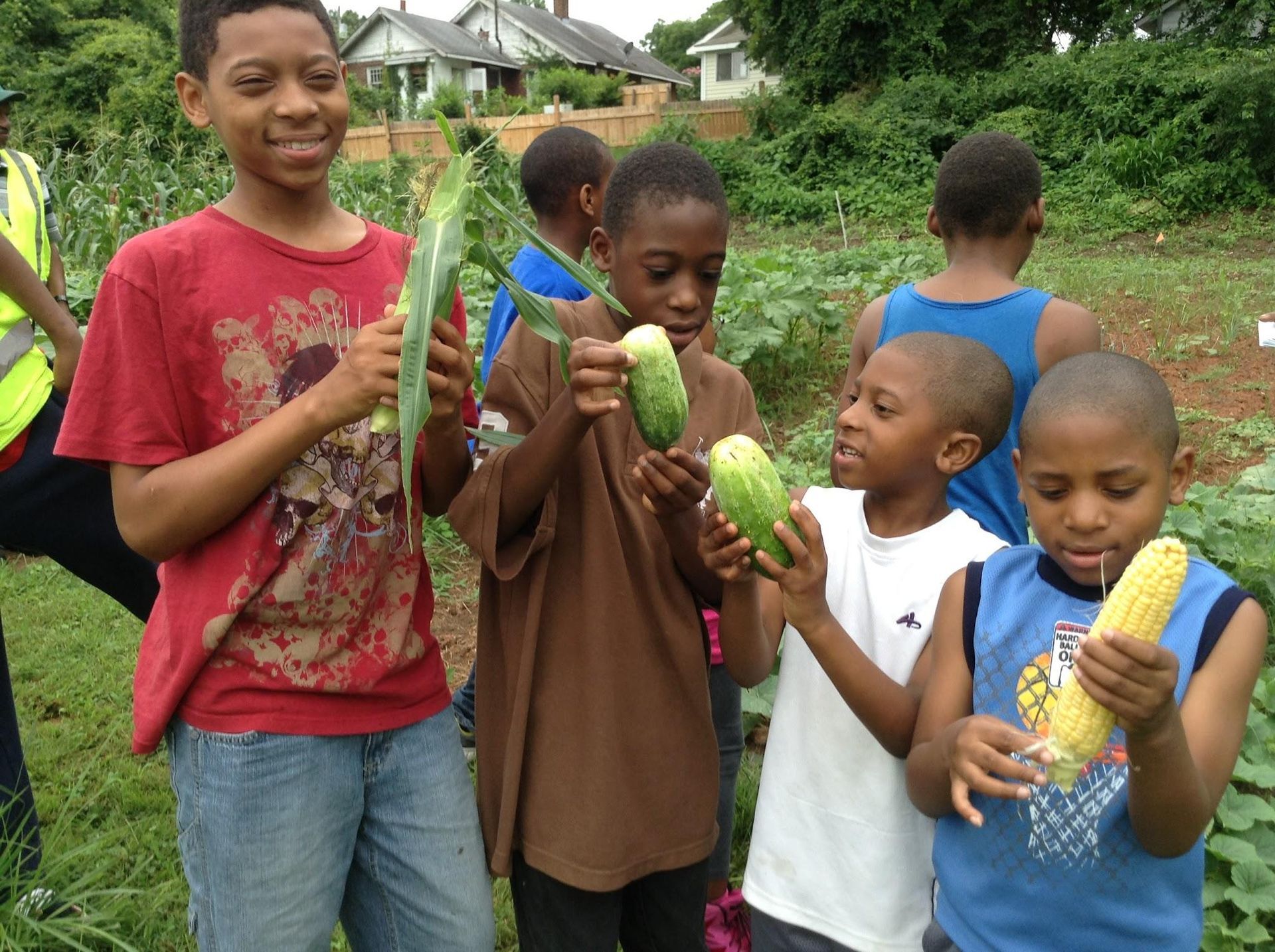 Five children holding fresh corn and cucumbers while standing in a community garden.