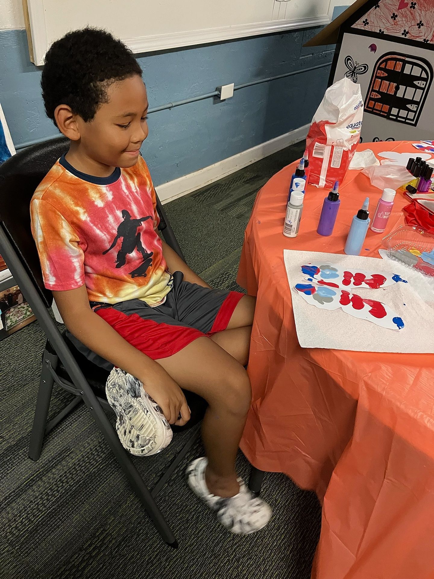 A person in a tie-dye shirt sits at an orange-covered table with art supplies and a craft project.