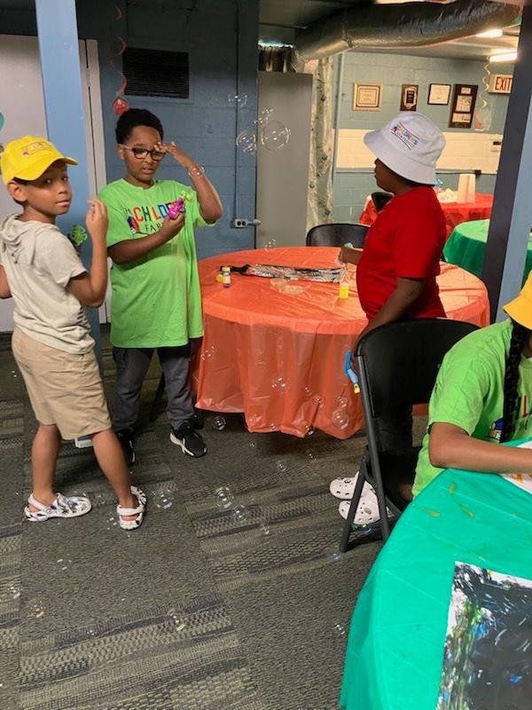 Children in colorful shirts and hats blow bubbles at round tables with orange and green tablecloths in an indoor space.