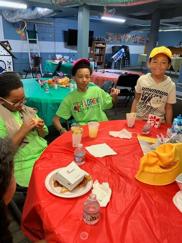 Three children sit at a round, red-covered table in a room with a green-covered table, eating Chick-fil-A meals.