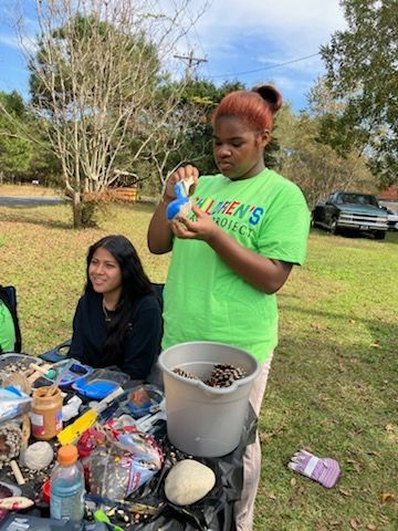 Two people crafting outdoors at a table with paint and pinecones, one holding a painted object in front of a bucket.