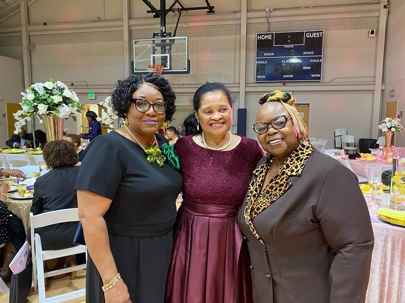 Three people pose for a photo in a gymnasium decorated for an event, smiling at the camera.
