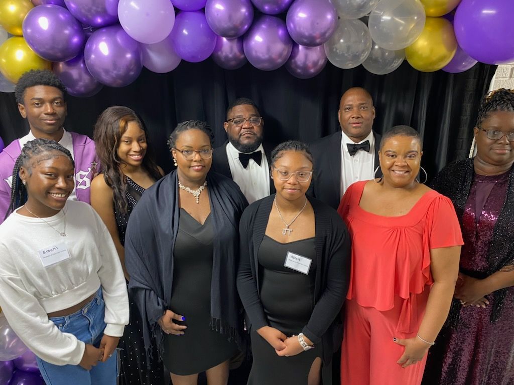 A group posing in front of a black backdrop decorated with gold and purple balloons at an event.
