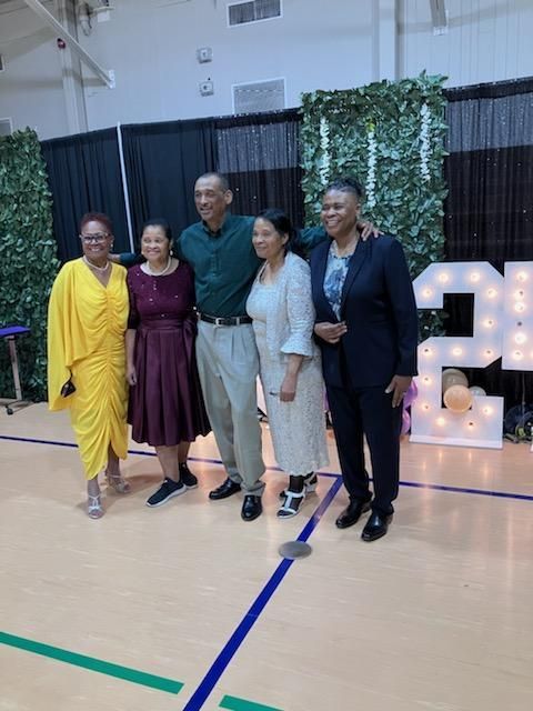 Five people stand smiling for a photo indoors in front of a green wall and a large, lighted number 24 display.