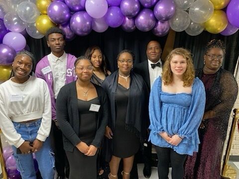 A group of seven people stand posed for a photo in front of a purple and gold balloon arch at an indoor event.