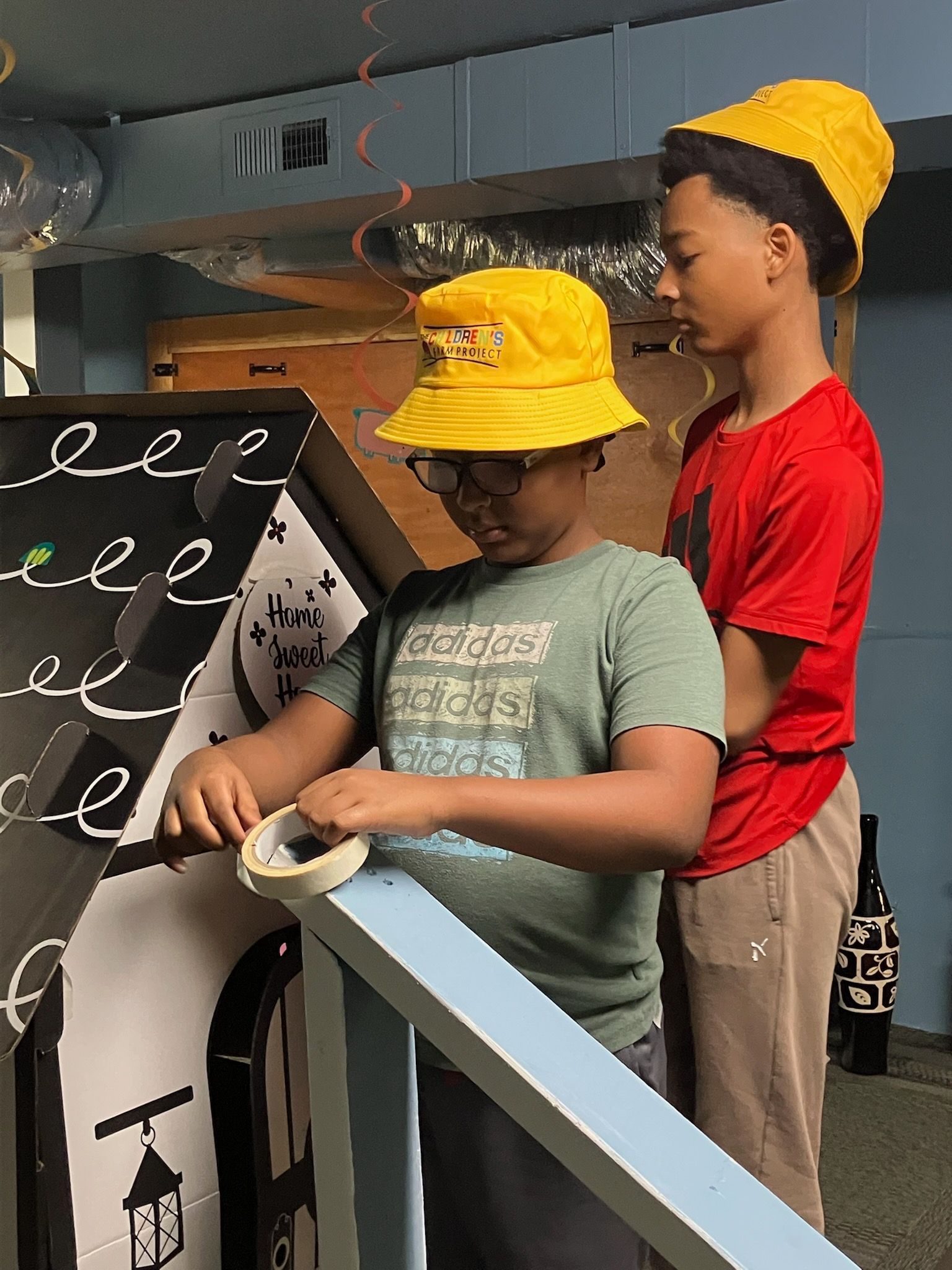 Two people wearing yellow bucket hats work together on a craft project using tape on a large, decorated cardboard structure.