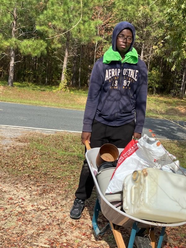 A person in a hooded sweatshirt and bright green neck gaiter stands beside a wheelbarrow filled with assorted supplies.
