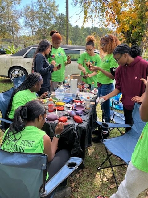 A group of people sit around a table outdoors, painting rocks with bright colors during a sunny daytime event.