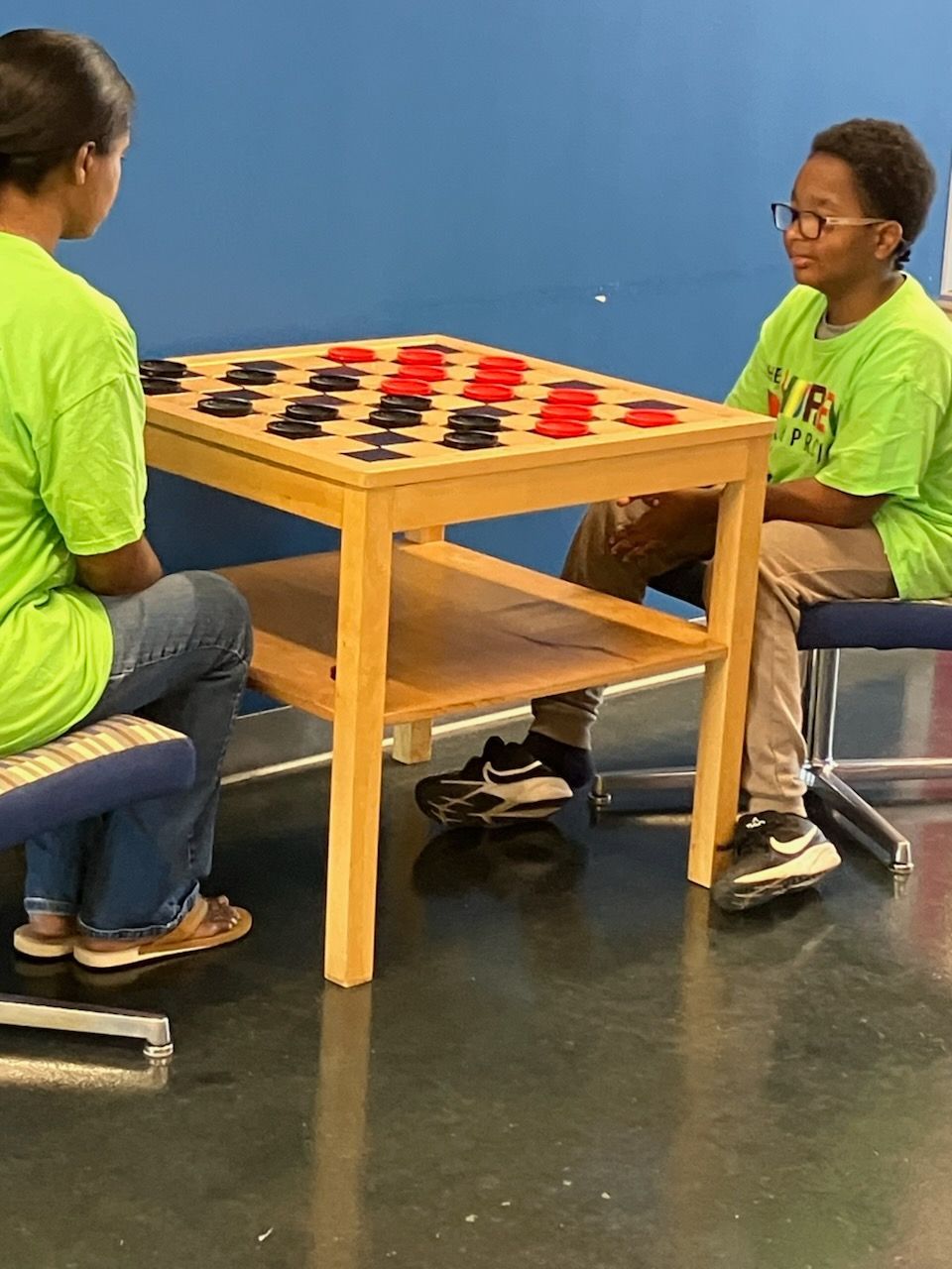 Two people in matching green shirts sit across from each other at a wooden table playing a game of checkers.