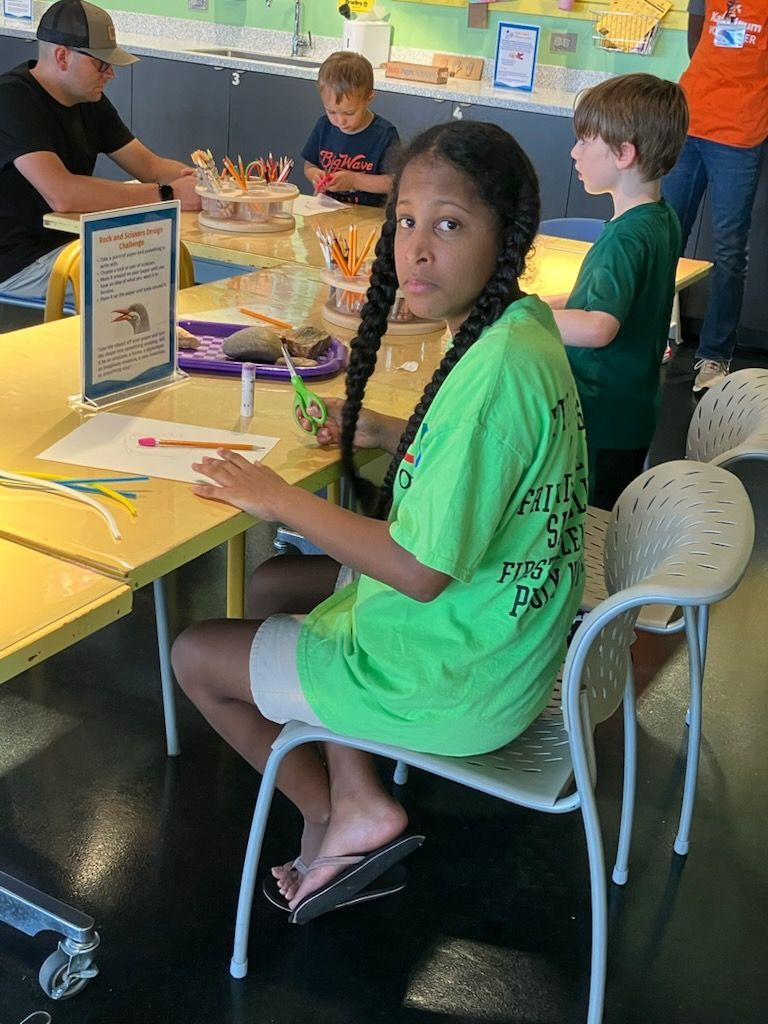 A person in a neon green shirt sits at a craft table with others, working on a project with colorful items.