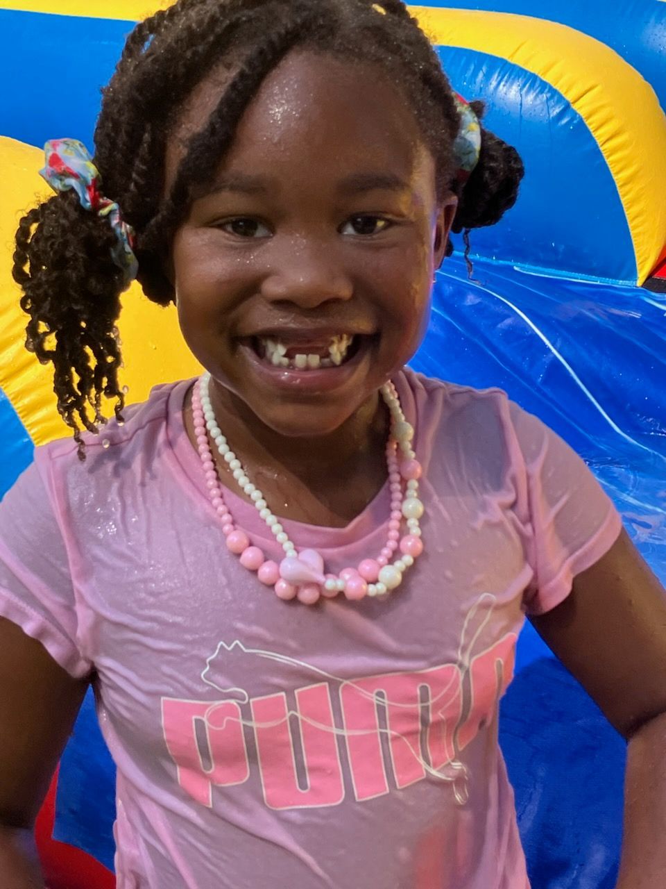 A smiling child in a pink t-shirt and beaded necklace standing in front of a blue and yellow inflatable slide.
