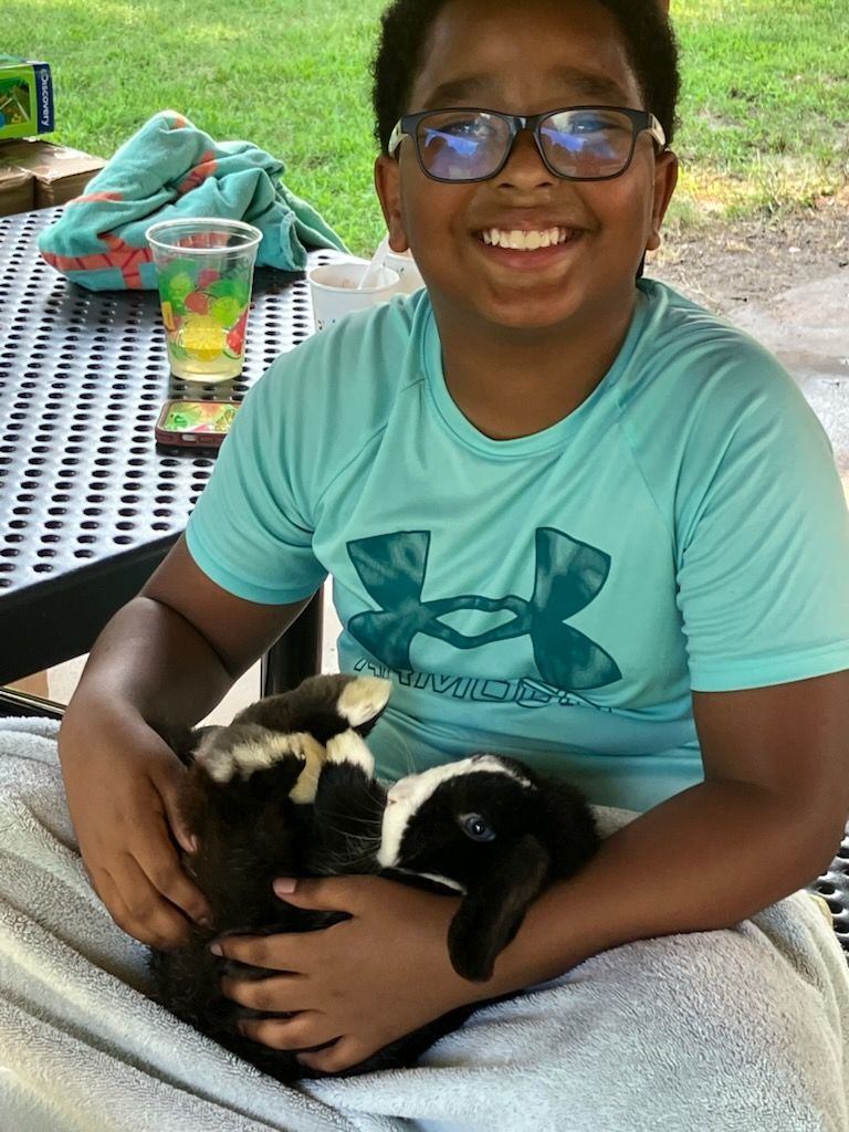 A smiling person in a teal shirt holds a small black and white rabbit while sitting at a table outdoors.
