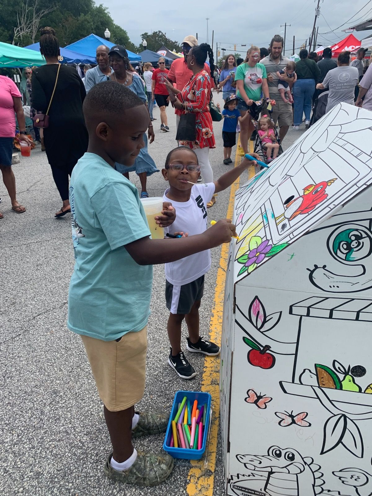 Two children color a large outdoor mural with markers at a sunny street festival with many people in the background.