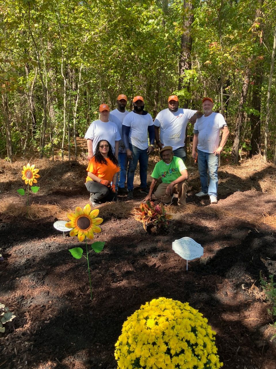 A group stands in a wooded area with freshly mulched ground, decorative sunflowers, and yellow mums in the foreground.