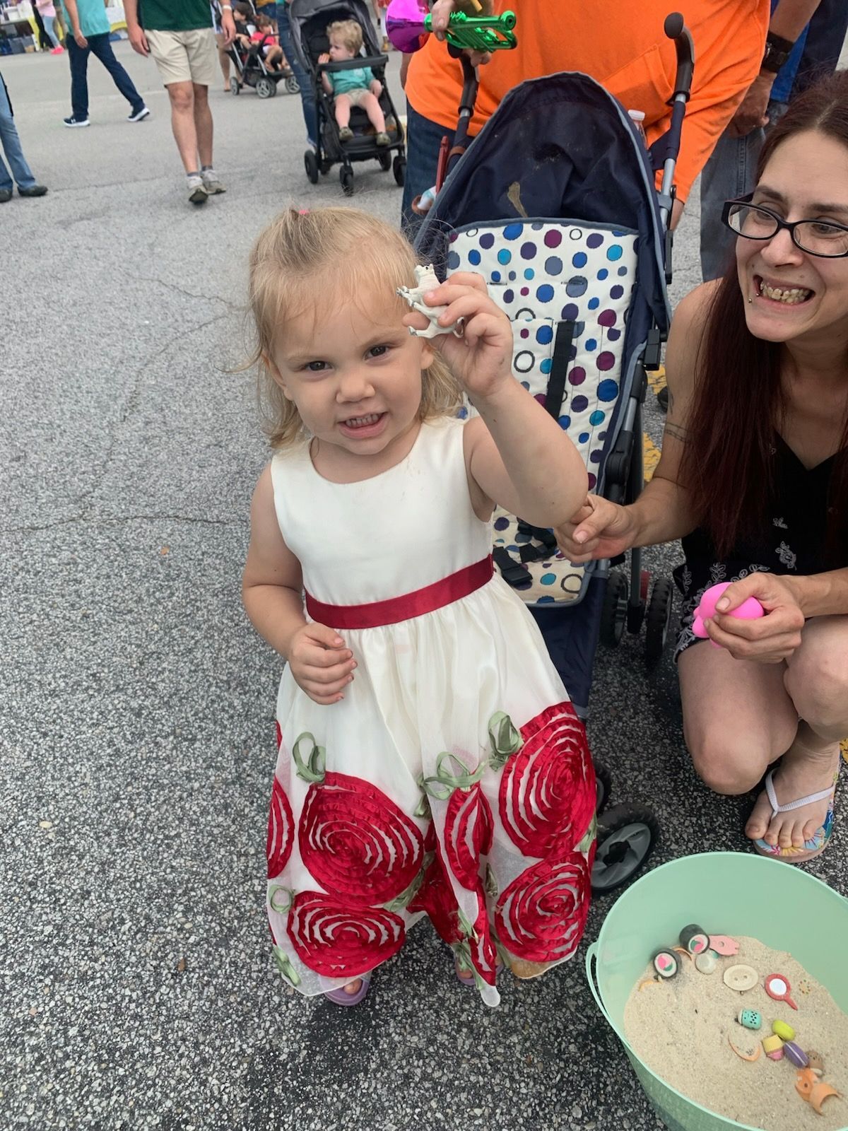 A toddler in a floral white dress holds a toy, standing near a woman and a stroller filled with sand and toys.