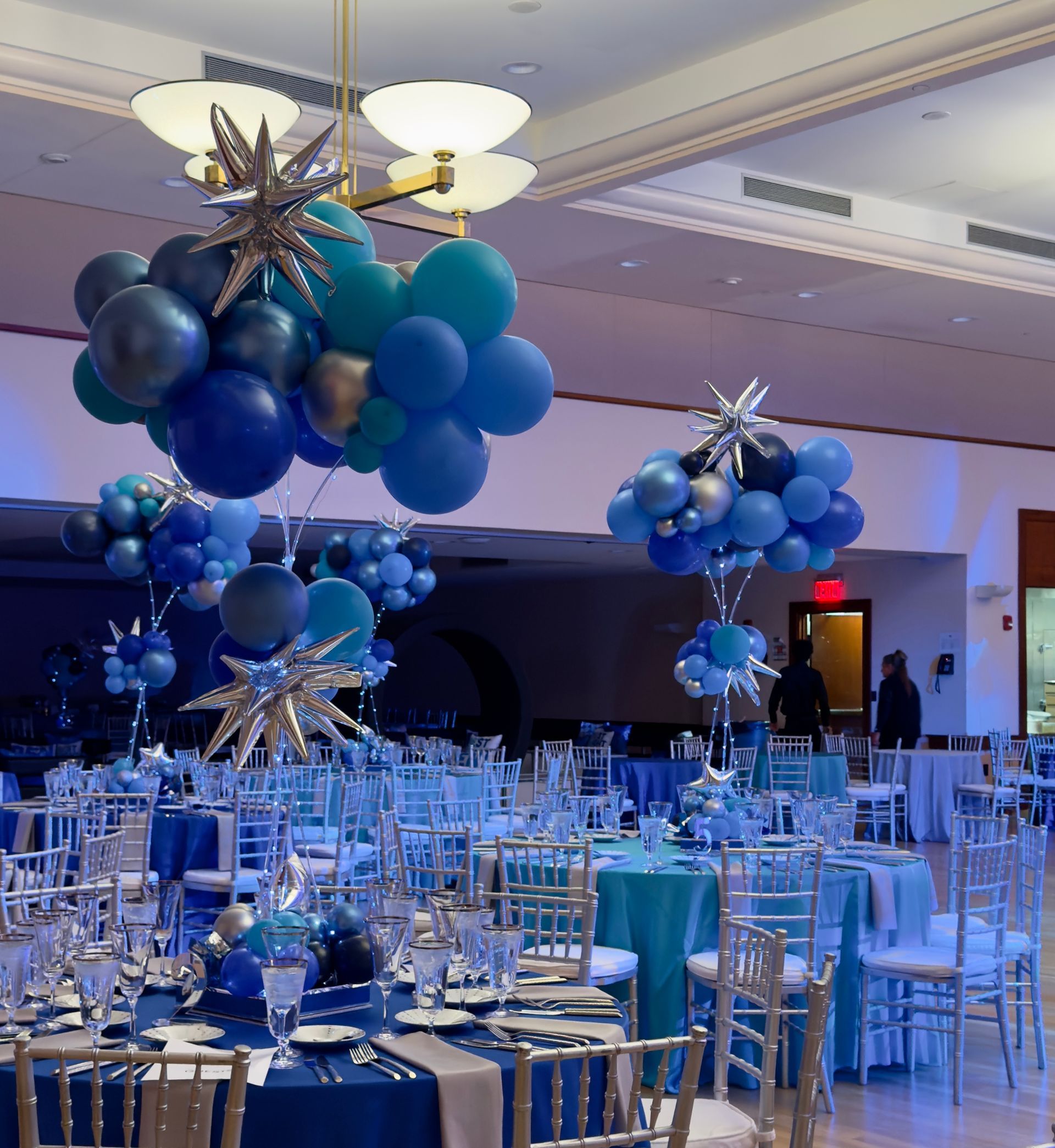 Blue and silver balloon arrangements decorate a banquet hall with tables set for a Bar Mitzvah