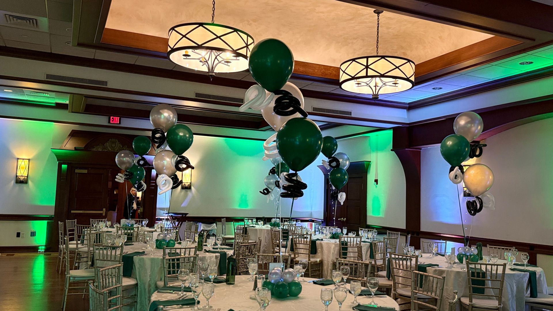 Mitzvah venue with green, navy blue, and silver balloons. Tables set, ambient lighting.