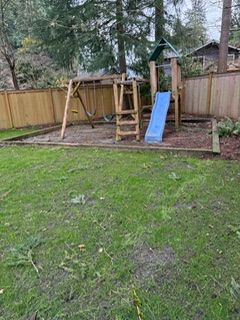 A wooden swing set and play structure with a blue slide sit in a backyard enclosed by a wooden fence.
