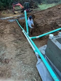 A construction worker stands in a trench next to green PVC drainage pipes near a concrete foundation and an excavator.