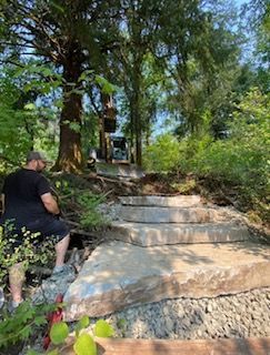 A person sits near large, newly installed stone steps on a wooded slope while heavy machinery works in the background.