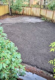 A freshly leveled, dark soil patch in a backyard, enclosed by a wooden fence and bordered by lush green bushes.