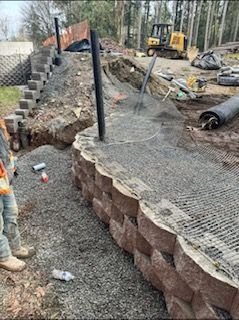 Construction worker stands near a curved segmental retaining wall with backfill, geogrid, and heavy machinery in the background.