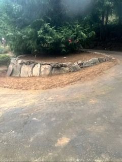 A crescent-shaped stone retaining wall borders a garden bed with a large evergreen bush, set beside an asphalt driveway.
