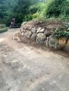 A rock retaining wall borders a dirt driveway with a small construction vehicle parked in the background.
