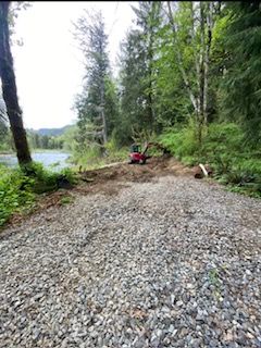 A small red excavator sits on a dirt path next to a riverbank in a wooded area, with gravel in the foreground.