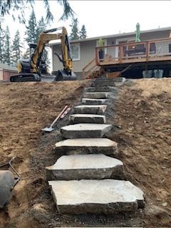 A series of large, irregular stone steps leading up a dirt hill toward a residential deck with an excavator parked nearby.