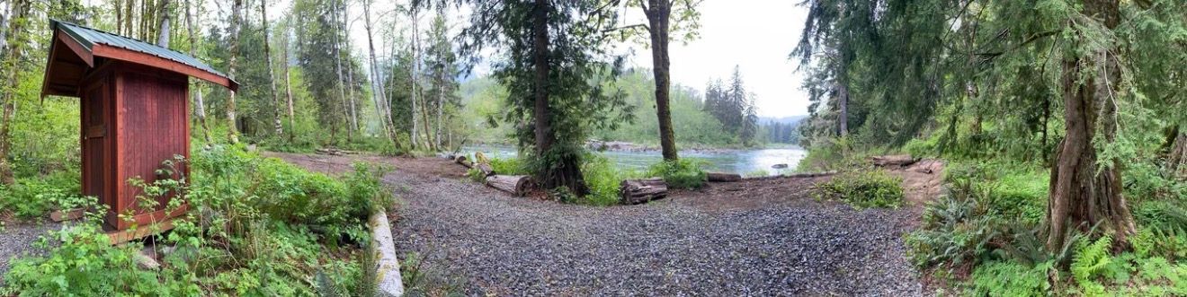 A wooden outhouse stands next to a gravel path leading to a lake surrounded by forest.