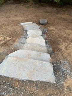 A path of gray stone slabs leading uphill, set into a dirt slope with gravel edges in an outdoor, wooded area.