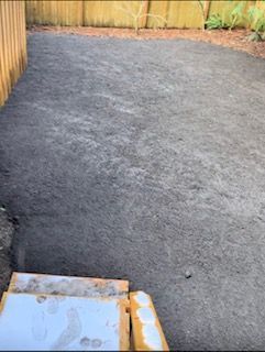 A view of a backyard featuring a freshly leveled, dark soil patch next to wooden stairs and a wooden fence.